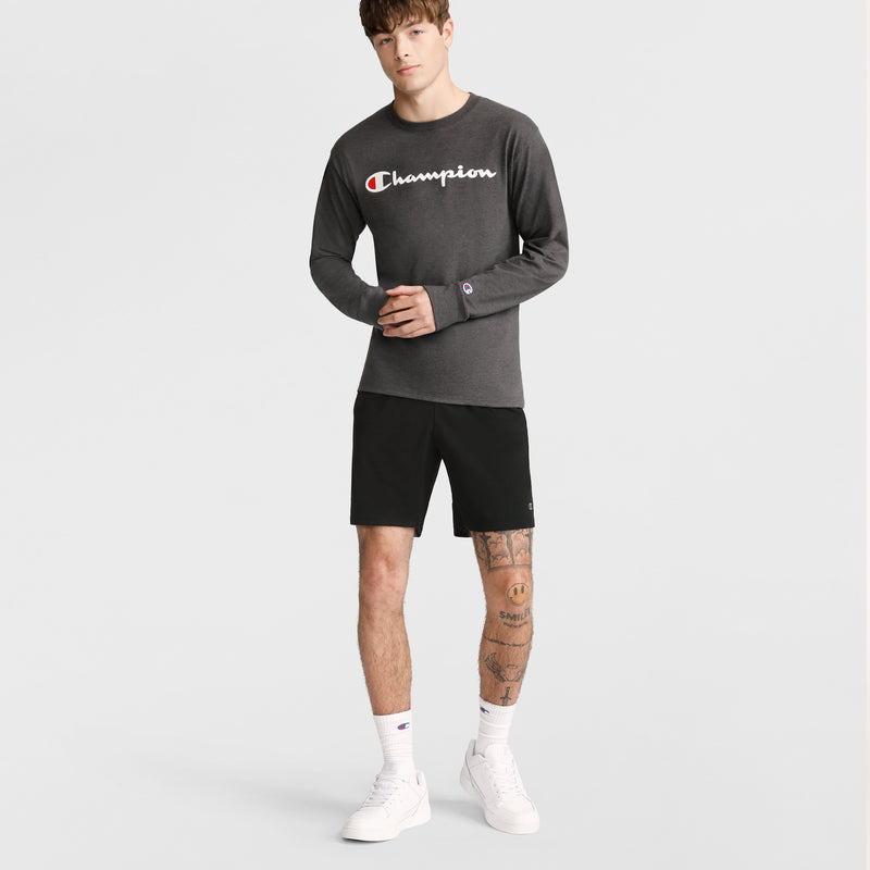 A man with tattoos on his left leg and brown, bang-styled hair stands against a plain background wearing a dark gray Champion long-sleeve shirt and Champion 10" Moisture Wicking Sport Shorts, paired with white socks and white sneakers.