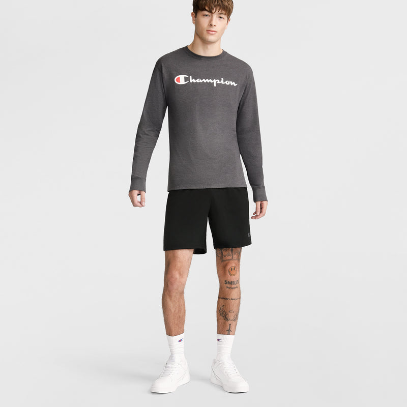 A young man with tattoos on his right leg wears Champion 10" Moisture Wicking Sport Shorts, a gray long-sleeve Champion shirt, white socks, and white sneakers against a plain background, showing a neutral expression.