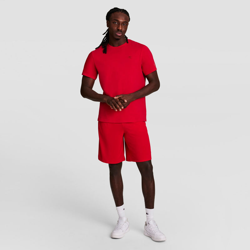 A man with long braided hair stands against a plain light gray background, wearing a red t-shirt and Champion 10" Moisture Wicking Sport Shorts, paired with white socks and sneakers. His hands are clasped in front of him.