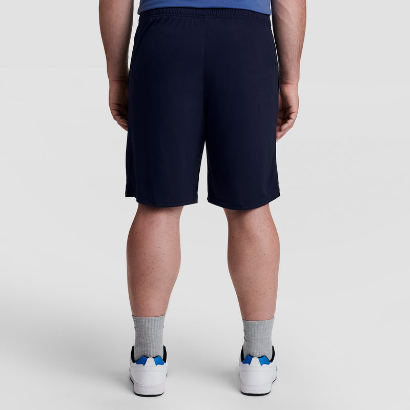 A person stands facing away from the camera, wearing Champion 10" Moisture Wicking Sport Shorts - Big & Tall in navy, gray socks, white sneakers with blue accents, and a blue shirt against a light plain background.