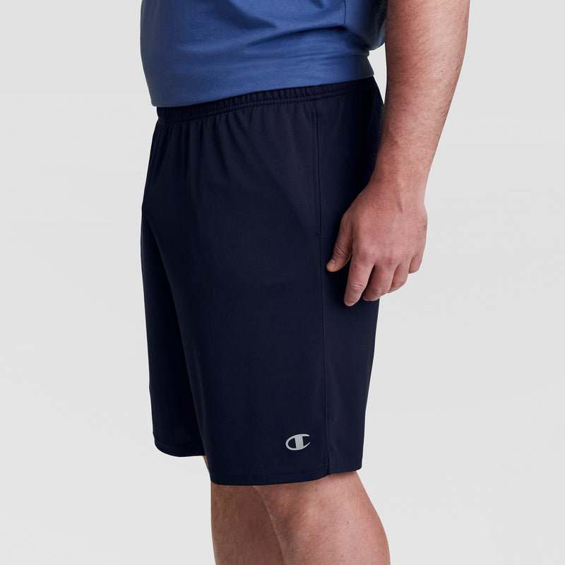 A person in a blue shirt and Champion 10" Moisture Wicking, Sport Shorts - Big & Tall stands facing right. The black shorts have an elastic waistband, small Champion logo near the hem, and moisture-wicking fabric. Plain light gray background.