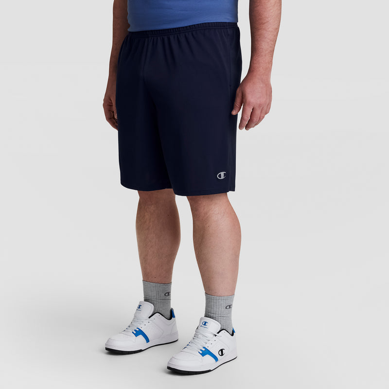 A person wearing a blue shirt, Champion 10" Moisture Wicking Sport Shorts - Big & Tall, gray Champion socks, and white sneakers with blue accents stands against a plain light background.