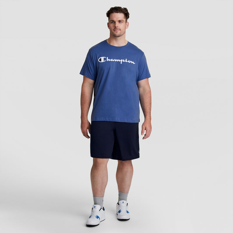 A man stands facing forward, wearing a blue Champion DOUBLE DRY t-shirt, Champion 10" Moisture Wicking Sport Shorts - Big & Tall in navy, gray socks, and white sneakers against a plain light gray background.