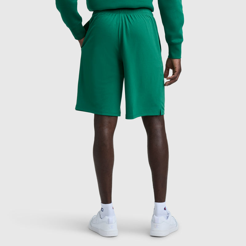 A person faces away, wearing a green sweatshirt, Champion 10" Moisture Wicking Sport Shorts with elastic waistband and side pockets, plus white sneakers and socks against a plain light background.