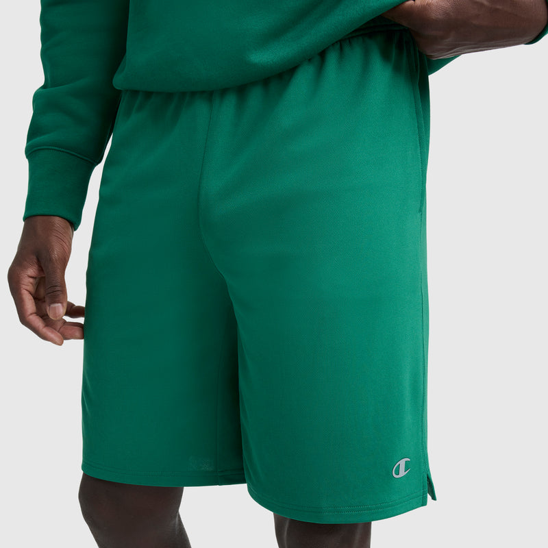 A person wearing a green long-sleeve shirt and Champion 10" Moisture Wicking Sport Shorts with an embroidered logo on the left leg stands against a plain, light background.