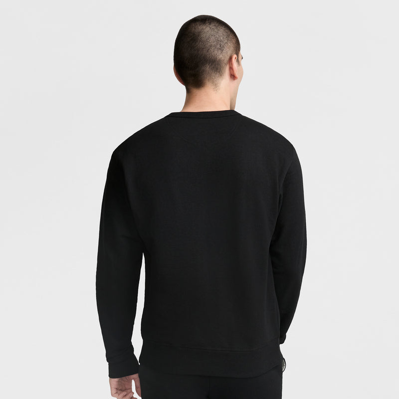 A person with short hair stands facing away from the camera against a plain light background, wearing a black Champion Powerblend Crewneck Sweatshirt with the Classic Script Logo.