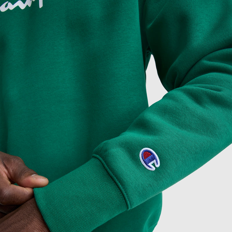 A person in a green Champion Powerblend Crewneck Sweatshirt, Classic Script Logo, adjusts the cuff with their other hand.