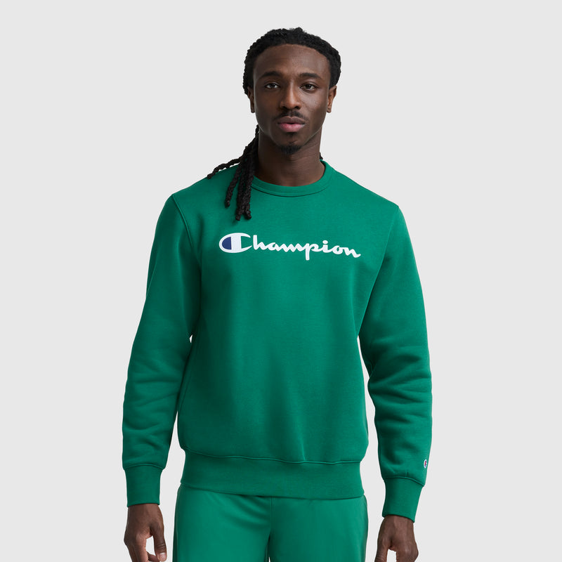A man with long braids wears a green Champion Powerblend Crewneck Sweatshirt with the classic script logo and matching green pants, standing against a plain light gray background.
