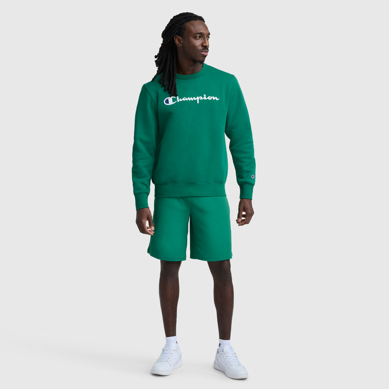A man stands against a plain background wearing a green Champion Powerblend Crewneck Sweatshirt, Classic Script Logo, with matching green shorts and white sneakers. He looks to his left with his arms relaxed by his sides.