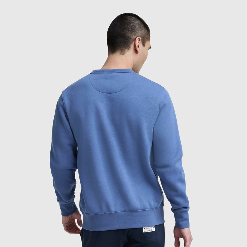 A person with short dark hair is seen from behind wearing a blue Champion Powerblend Crewneck Sweatshirt, Classic Script Logo, and dark pants against a plain light gray background.