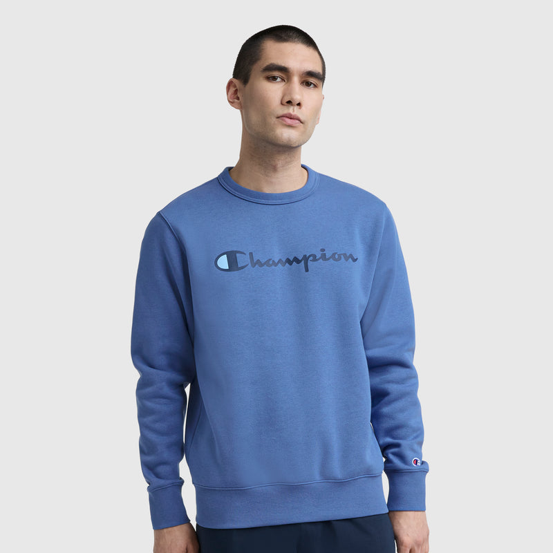 A man stands against a plain light background, wearing the Champion Powerblend Crewneck Sweatshirt with the Classic Script Logo across the chest, and looks directly at the camera with a neutral expression.