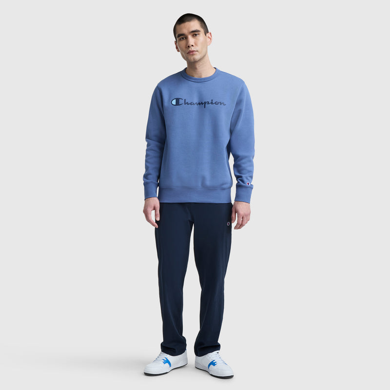 A man stands facing forward, wearing a Champion Powerblend Crewneck Sweatshirt with the classic script logo in blue, dark blue pants, and white sneakers, set against a plain light gray background.