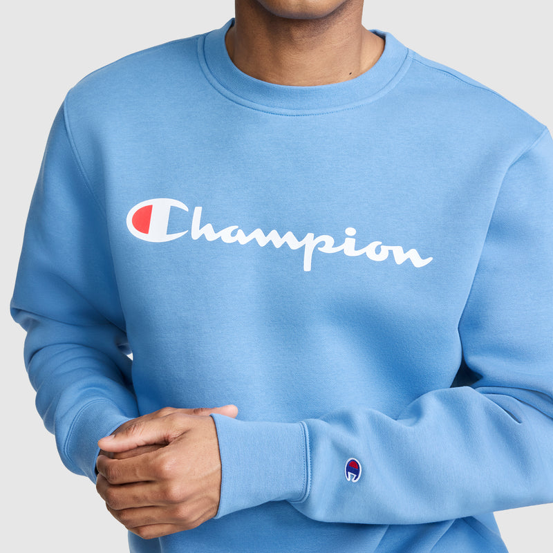A person is wearing the Champion Powerblend Crewneck Sweatshirt, Classic Script Logo—a light blue men’s fleece pullover featuring the iconic Champion logo in white and red across the chest and a small logo on the sleeve—hands clasped together. This versatile sweatshirt delivers comfort and style for everyday wear.