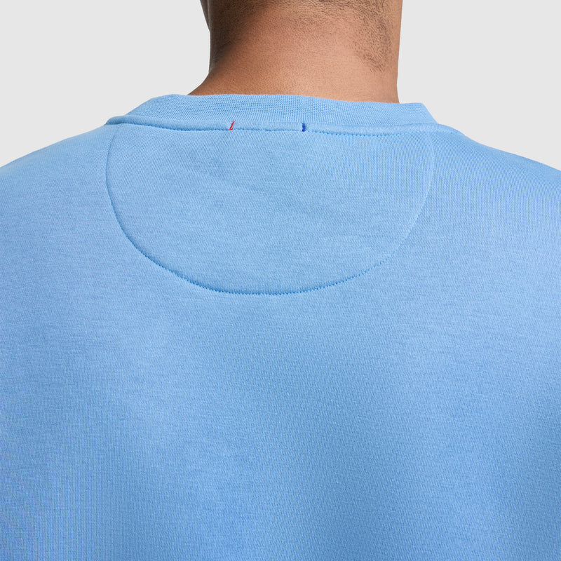 A close-up shows the back of a person’s neck and shoulders, featuring the light blue Champion Powerblend Crewneck Sweatshirt with Classic Script Logo. Visible stitching and a circular patch accent the upper back area, and the person’s hairline is also visible.