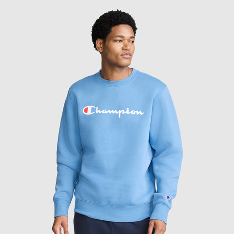 A person stands against a plain gray background, looking to the side with a neutral expression while wearing the Champion Powerblend Crewneck Sweatshirt, Classic Script Logo in light blue. The iconic Champion logo is printed across the chest.