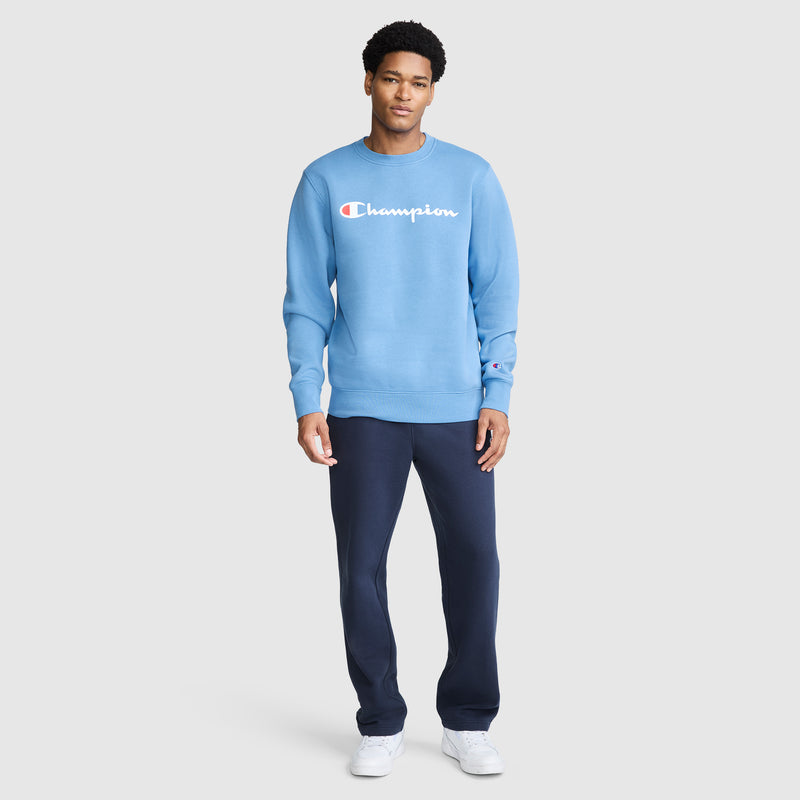 A man stands facing forward, wearing a light blue Champion Powerblend Crewneck Sweatshirt with Classic Script Logo, dark blue pants, and white sneakers, against a plain light gray background.