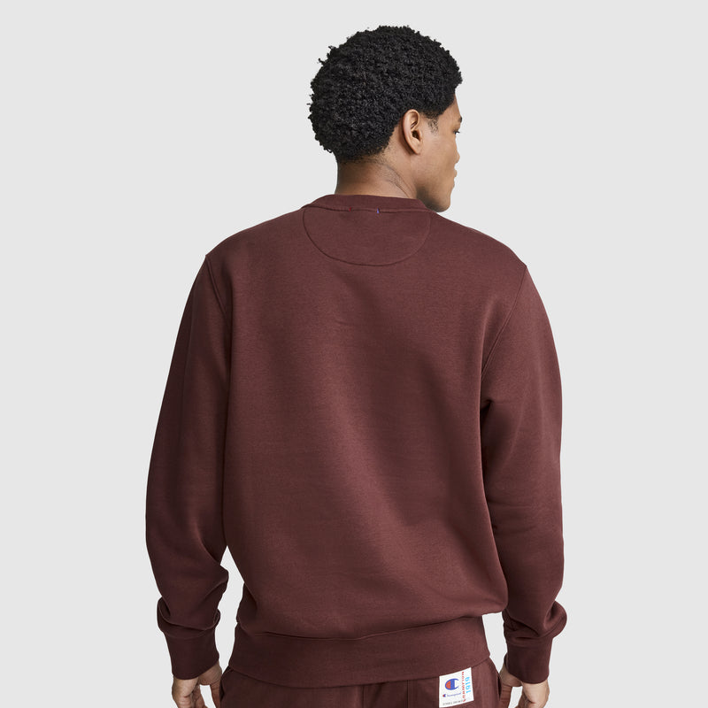 A person wearing a maroon Champion Powerblend Crewneck Sweatshirt with the Classic Script Logo and matching pants stands facing away, displaying the back of the outfit against a plain light gray background.