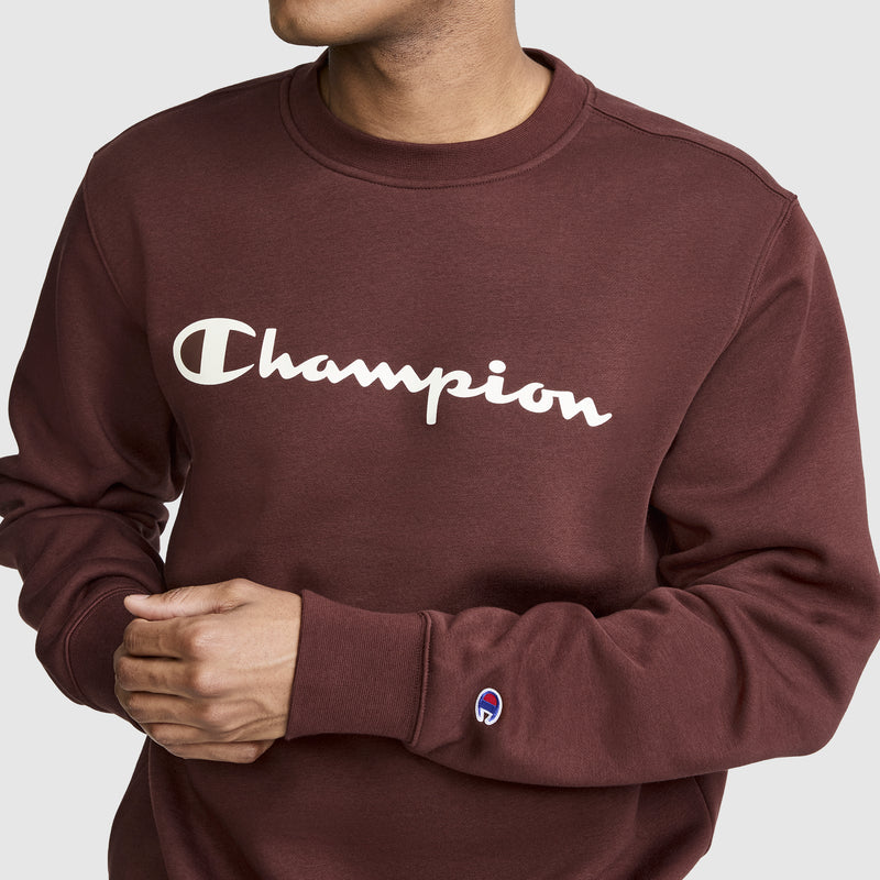 A person wearing a maroon Champion Powerblend Crewneck Sweatshirt, Classic Script Logo stands against a plain background. Only their upper body is visible, with their head partially out of frame. The crewneck features the signature Champion script logo printed across the chest.