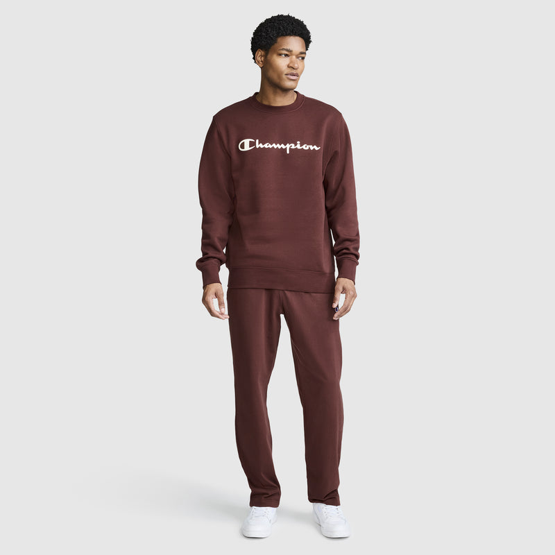A person stands against a plain white background, looking to the side, dressed in a maroon Champion Powerblend Crewneck Sweatshirt with the classic script logo and matching pants. They are also wearing white sneakers, highlighting the comfort and timeless style of this Champion sweatshirt.