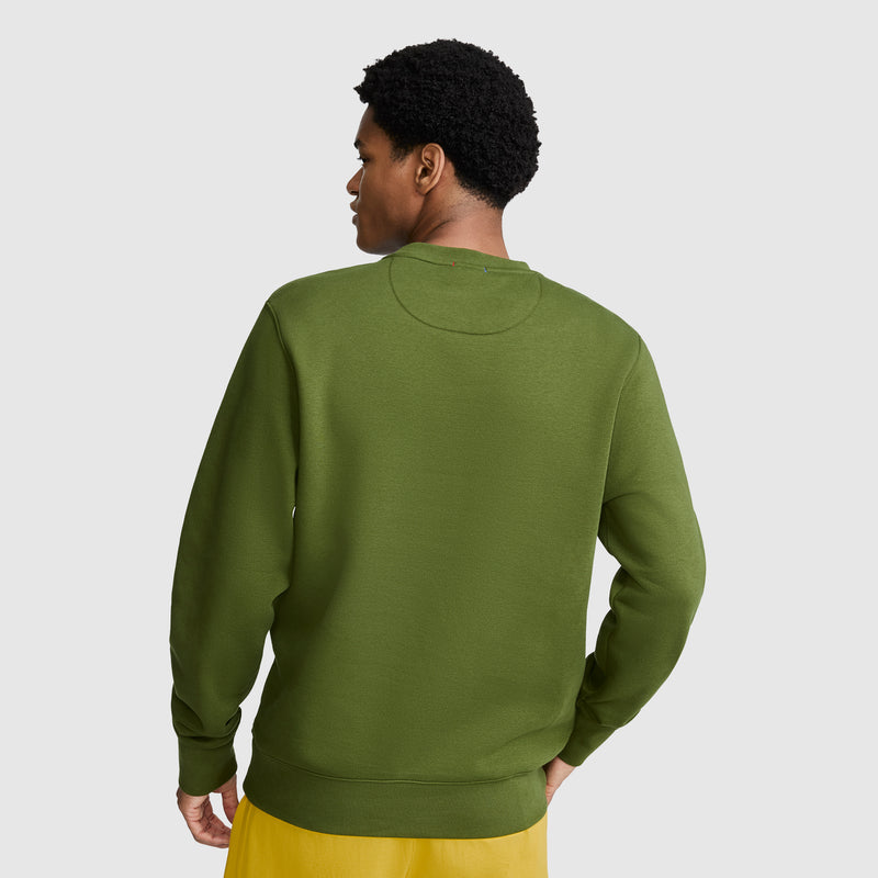 A person stands facing away, wearing a green Champion Powerblend Crewneck Sweatshirt with the Classic Script Logo and yellow shorts against a plain light gray background.