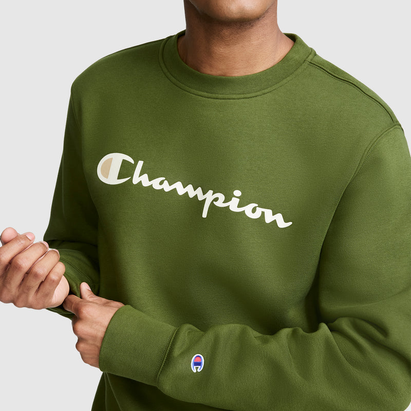 A person wearing a Champion Powerblend Crewneck Sweatshirt, Classic Script Logo, in olive green, featuring the iconic Champion script logo across the chest and a small blue, red, and white patch on the sleeve. This fleece sweatshirt offers both comfort and style with its classic crewneck Powerblend design.