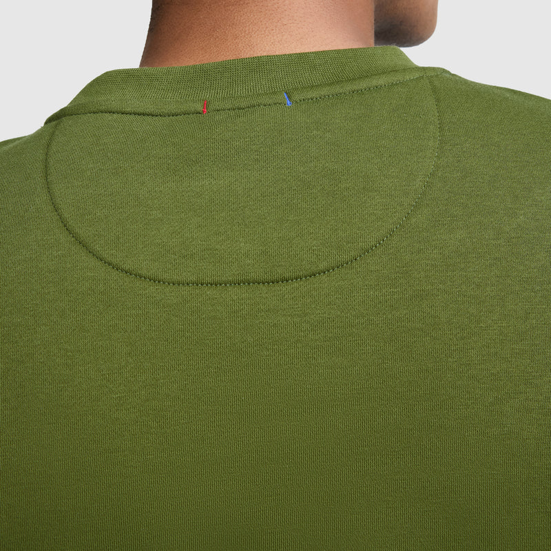 Close-up of the back of a person’s neck wearing a green Champion Powerblend Crewneck Sweatshirt, Classic Script Logo, highlighting the detailed stitching and small red and blue thread accents near the collar.