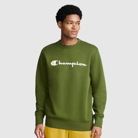 A man wearing a green Champion Powerblend Crewneck Sweatshirt with the Classic Script Logo and yellow shorts stands against a plain light gray background, looking to his left with a neutral expression.