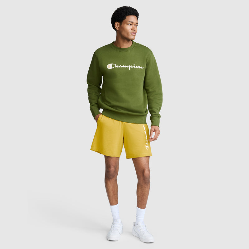 A man wearing a green Champion Powerblend Crewneck Sweatshirt with the Classic Script Logo, yellow Champion shorts, white socks, and white sneakers stands against a plain light gray background.