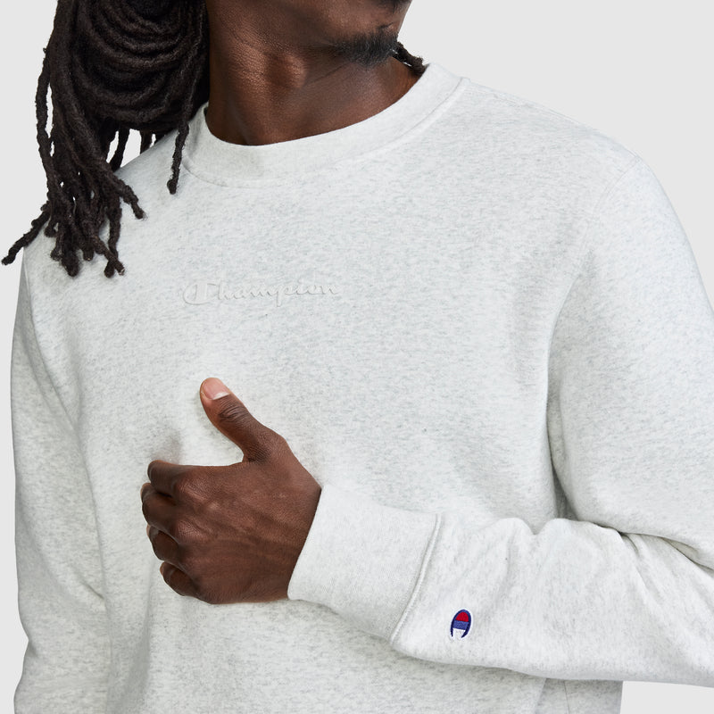 A person with long dreadlocks wears a light gray Champion Powerblend Crewneck Sweatshirt featuring a tonal script logo embroidered on the chest, and lifts one hand near their collar. The signature Champion patch is visible on the sleeve.