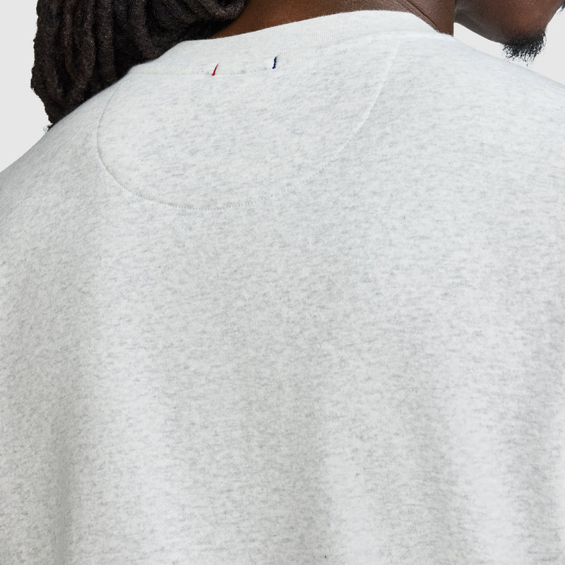 Close-up of the back of a person’s light gray Champion Powerblend Crewneck Sweatshirt, Tonal Script Logo, highlighting the upper back and neck area. A small red, white, and blue stitched Champion detail appears near the neckline. Part of the person’s face and hair is visible.