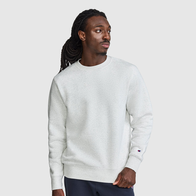 A man with long hair is wearing a Champion Powerblend Crewneck Sweatshirt with a tonal script logo in light gray and dark pants, standing against a plain white background and looking to his left with a neutral expression.
