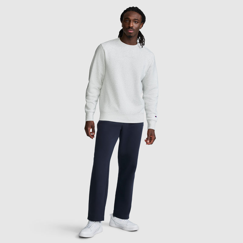 A man stands facing forward, wearing a Champion Powerblend Crewneck Sweatshirt with a tonal script logo in light gray, paired with dark blue pants and white sneakers. He has long braided hair and a neutral expression against a plain light background.