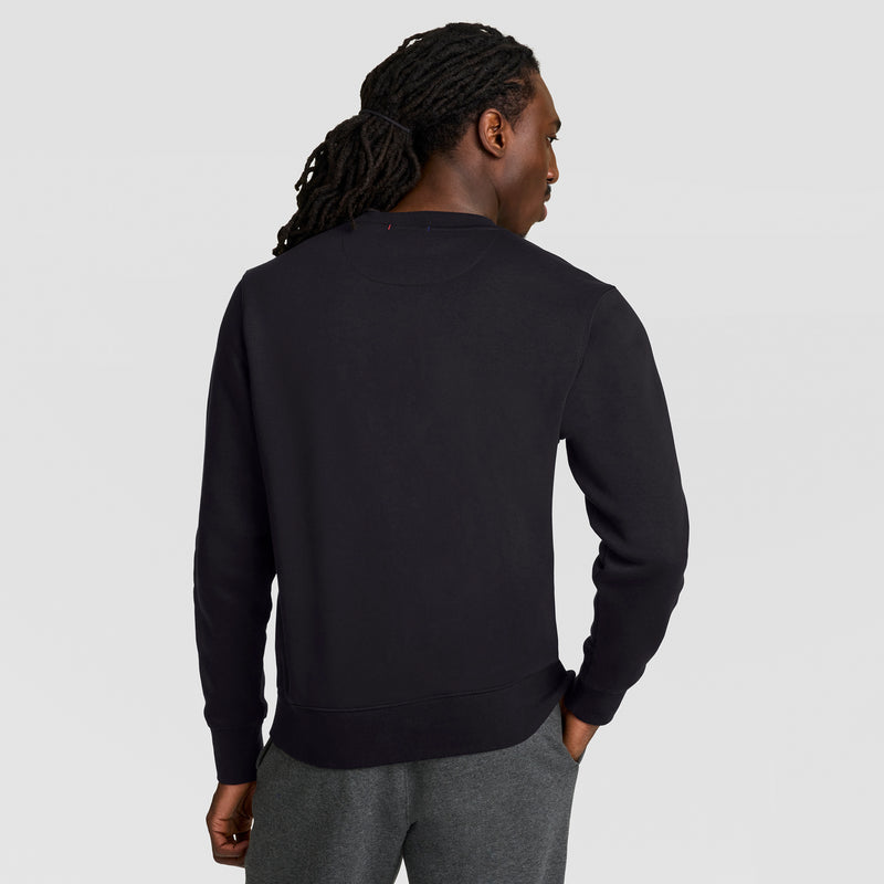 A person with long, braided hair stands with their back to the camera, wearing a black Champion Powerblend Crewneck Sweatshirt, Tonal Script Logo, and gray pants against a plain light gray background.