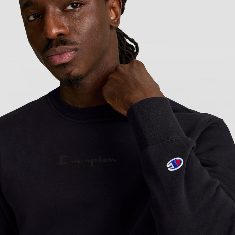 A man wears a black Champion Powerblend Crewneck Sweatshirt, Tonal Script Logo, featuring the brand’s logo on the chest and a small sleeve patch, made from soft Powerblend Fleece, standing against a plain light background.