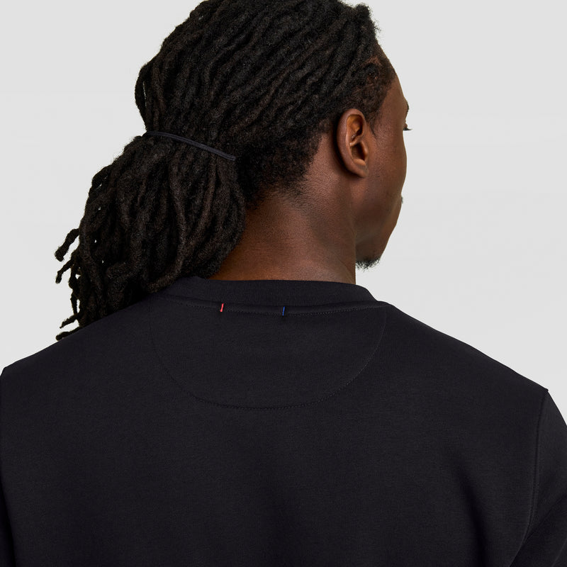 A person with long, dark dreadlocks tied back is seen from behind wearing a black Champion Powerblend Crewneck Sweatshirt, Tonal Script Logo, against a plain light background.