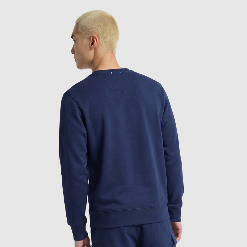 A person with short, bleached blonde hair is facing away from the camera, wearing a Champion Powerblend Crewneck Sweatshirt with Tonal Script Logo in navy blue and matching pants, set against a plain light gray background.