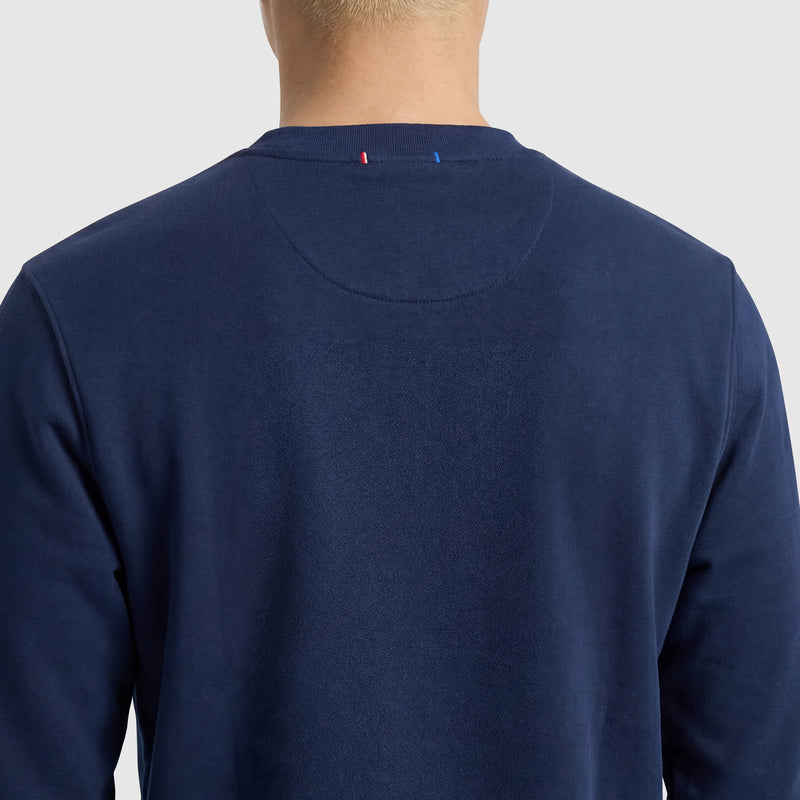 A person with short blonde hair is seen from behind wearing a Champion Powerblend Crewneck Sweatshirt, Tonal Script Logo in dark blue. The plain light gray background and small red, white, and blue stitched neckline details are visible.