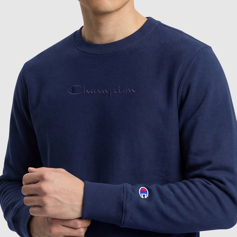 A person wears a navy blue Champion Powerblend Crewneck Sweatshirt, Tonal Script Logo, featuring an embroidered logo on the chest and a small patch on the left sleeve, standing against a light gray background.