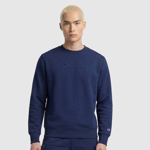 A young man with short blond hair wears a navy blue Champion Powerblend Crewneck Sweatshirt with Tonal Script Logo and matching pants, standing against a plain light gray background.