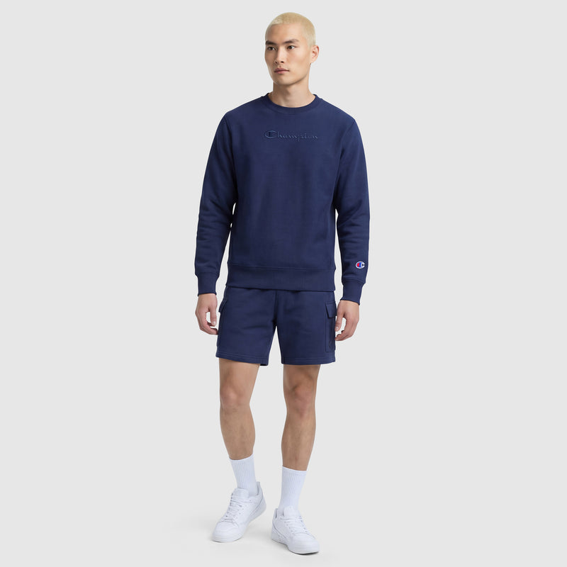 A person with short blond hair wears a Champion Powerblend Crewneck Sweatshirt, Tonal Script Logo, in navy blue with matching shorts, white socks, and white sneakers, standing against a plain light gray background.