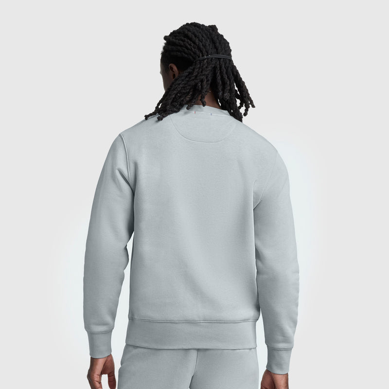 A person with long, twisted hair wears a Champion Powerblend Crewneck, Tonal Script Logo sweatshirt and matching sweatpants, standing with their back to the camera against a plain light background.