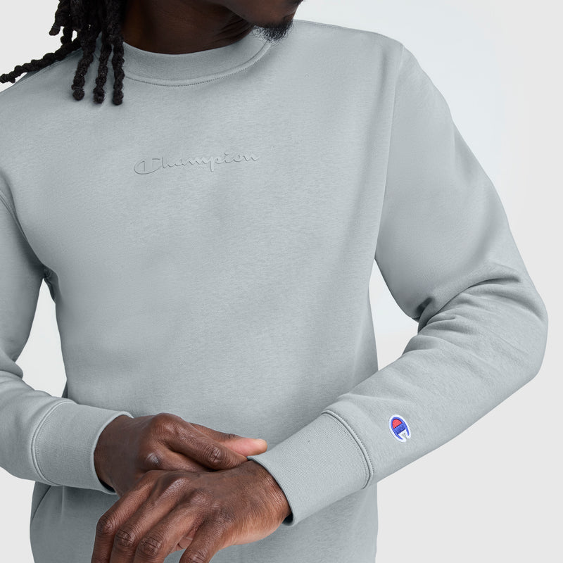 A person wearing a light gray Champion Powerblend Crewneck with a tonal script logo adjusts their sleeve. The men’s sweatshirt features an embroidered chest logo, cuff patch, and is made from soft Powerblend fleece for comfort.