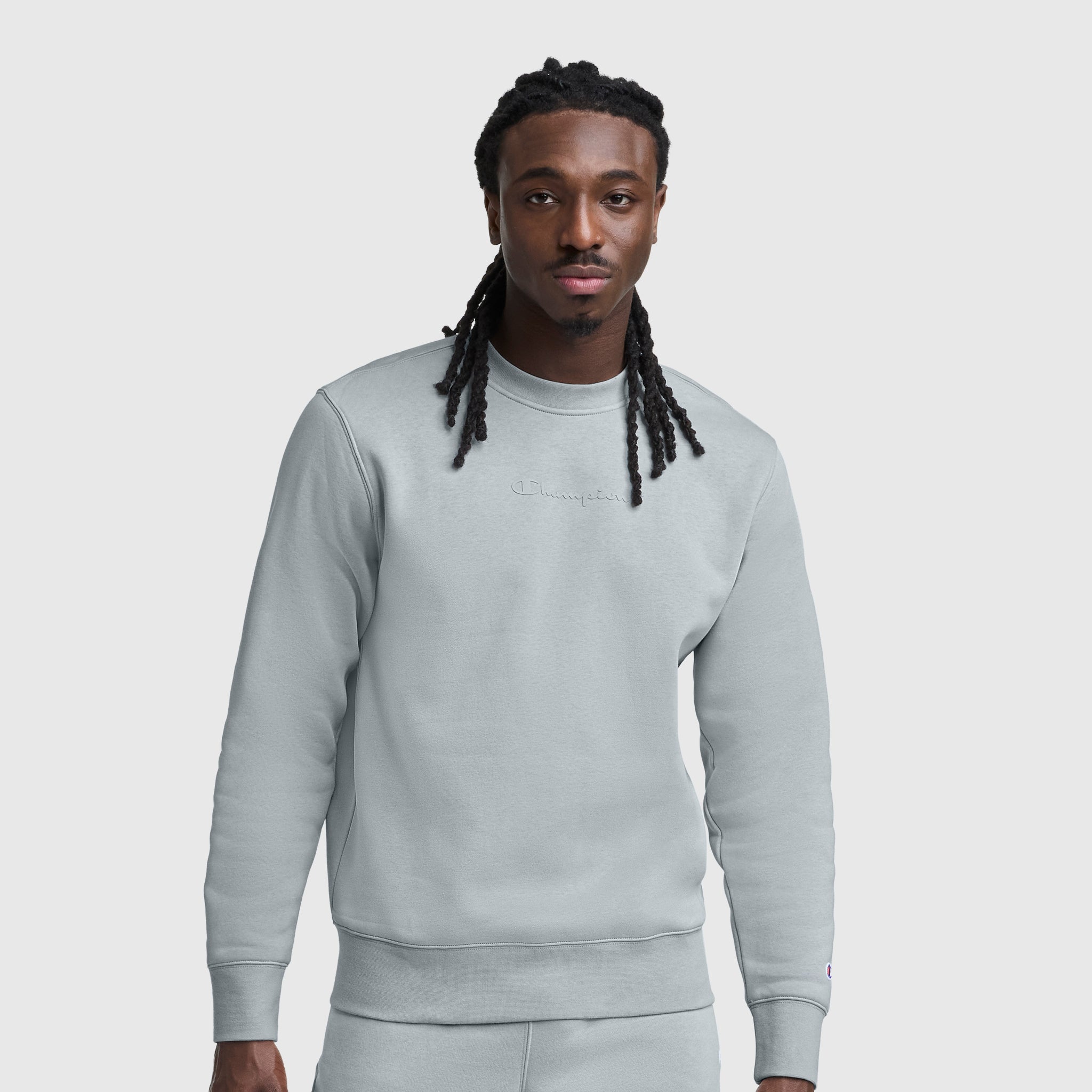 A man with long braids wears a Champion Powerblend Crewneck, Tonal Script Logo sweatshirt and matching pants, standing against a plain light background and looking at the camera with a neutral expression.