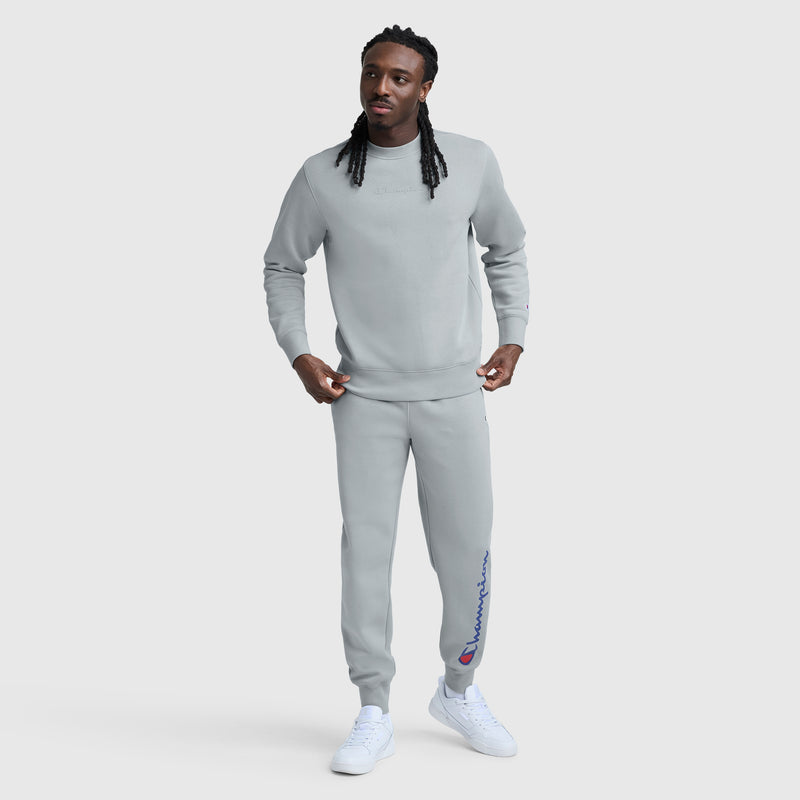A man with long braids wears a Champion Powerblend Crewneck with a tonal script logo and matching sweatpants. He stands on a white background, wearing white sneakers and looking slightly to the side.