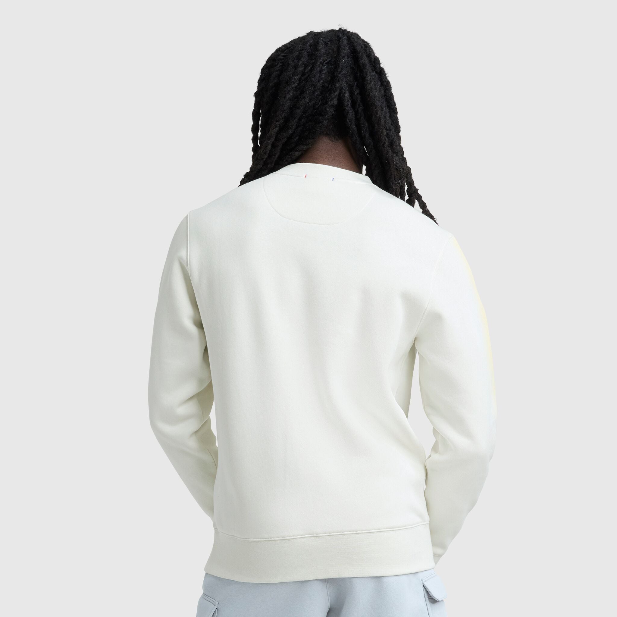 A person with long, dark, braided hair is seen from behind, wearing the Champion Powerblend Crewneck with Tonal Script Logo and light-colored pants against a plain gray background.