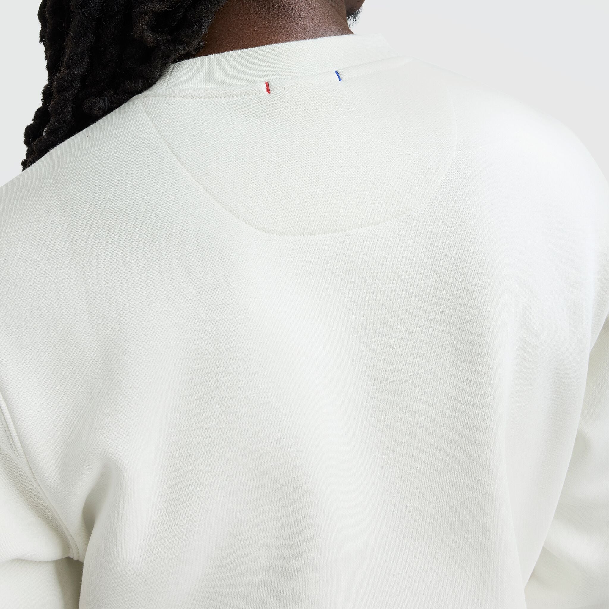 Close-up of a person wearing the Champion Powerblend Crewneck, Tonal Script Logo, seen from behind. The off-white fleece has subtle stitched red and blue details at the neckline. The person’s long, dark, twisted hair stands out against a light background.