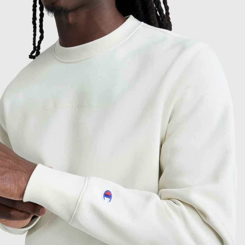 A person with dark skin and long braided hair wears a Champion Powerblend Crewneck, Tonal Script Logo, in off-white, featuring the brand's logo on the chest and a small sleeve patch, set against a plain light background.