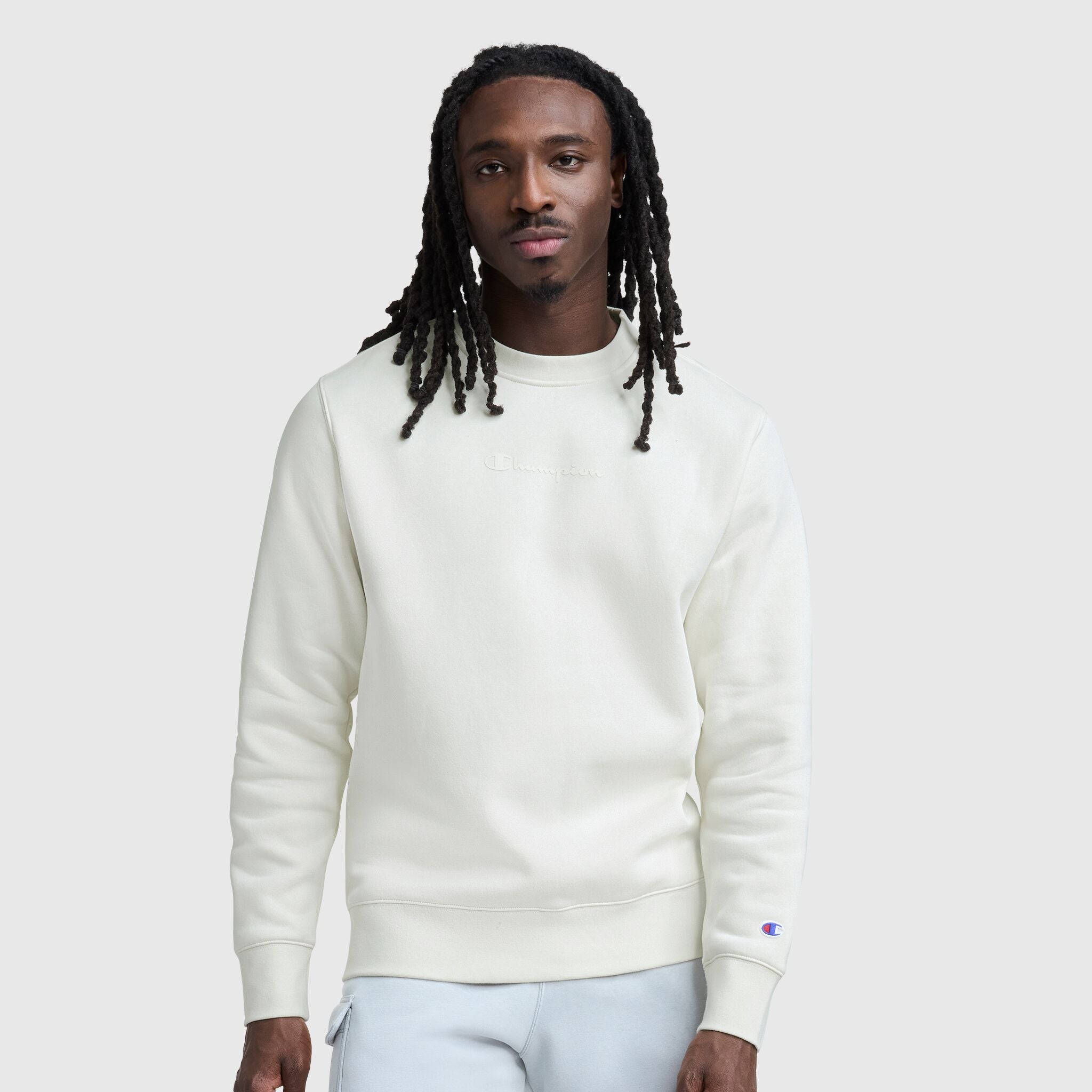 A man with long dreadlocks wears a cream Champion Powerblend Crewneck with a tonal script logo and light gray pants, standing against a plain light gray background and looking at the camera with a neutral expression.