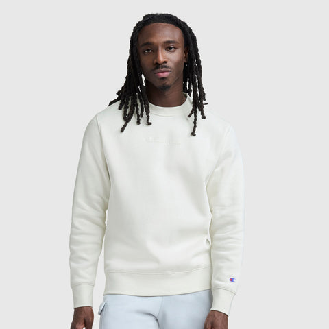 A man with long dreadlocks wears a cream Champion Powerblend Crewneck with a tonal script logo and light gray pants, standing against a plain light gray background and looking at the camera with a neutral expression.