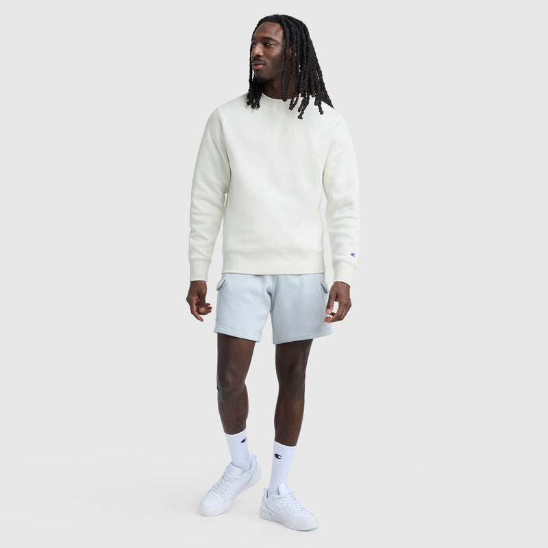 A man with long dreadlocks wears a Champion Powerblend Crewneck, Tonal Script Logo, paired with light gray shorts, white crew socks, and white sneakers, standing against a plain light gray background.